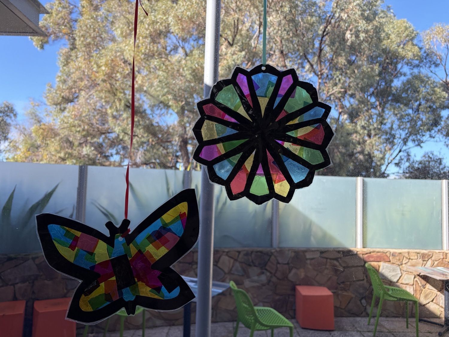 A colorful butterfly and a circular geometric mandala suncatcher hang from a window, looking out onto a sunny courtyard with green chairs